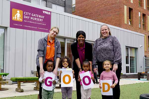 Children and staff happy and smiling, standing outside in their nursery garden holding a sign saying GOOD after their first successful OFSTED inspection.