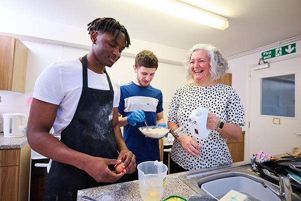 two young men and ymca housing support officer cooking together in communal kitchen, laughing and smiling.